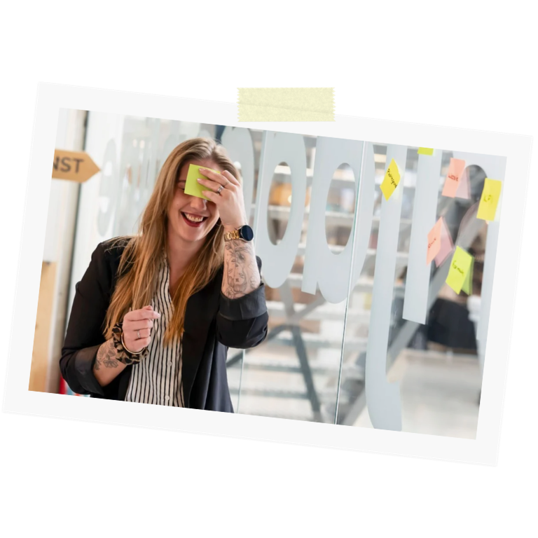 Tamara van Manager Happy lachend met een geel plakbriefje op haar voorhoofd voor een glazen wand met post-its