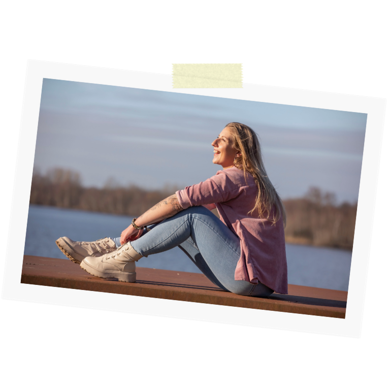 Een lachende jonge vrouw met blond haar zit buiten aan het water in het zonnetje